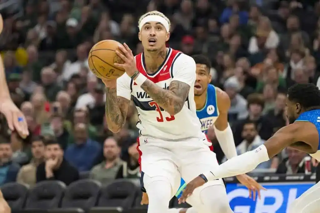 Washington Wizards player in a white jersey holding the ball mid-air, preparing for a shot while defended by Milwaukee Bucks players.