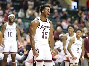 Texas A&M Aggies basketball player in a white uniform flexes and shouts in celebration while teammates cheer behind him.