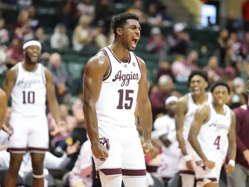 Texas A&M Aggies basketball player in a white uniform flexes and shouts in celebration while teammates cheer behind him.