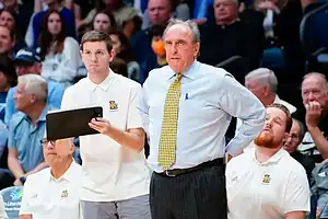 A basketball coach stands on the sidelines, instructing players and holding a clipboard, surrounded by team members in uniforms.