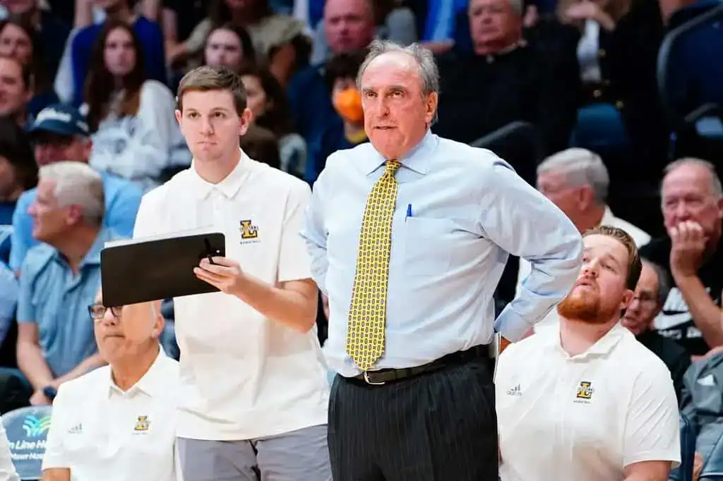 A basketball coach stands on the sidelines, instructing players and holding a clipboard, surrounded by team members in uniforms.