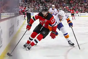 A New Jersey Devils player with the captain’s “C” skates along the boards while being closely pursued by a New York Islanders opponent.