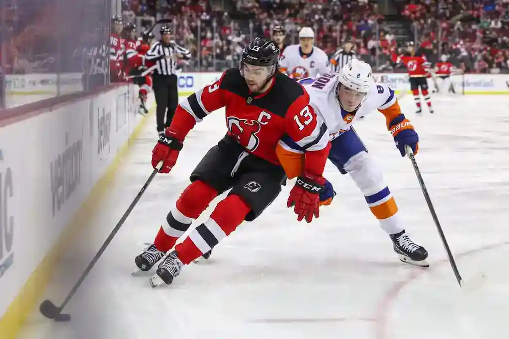 A New Jersey Devils player with the captain’s “C” skates along the boards while being closely pursued by a New York Islanders opponent.