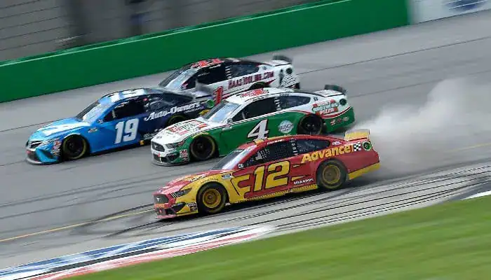 NASCAR cars race side by side at high speed, with one car skidding slightly and producing smoke.