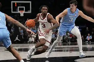 Oklahoma Sooners basketball player driving to the basket while being closely guarded by a North Carolina defender.