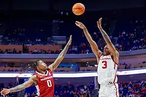 Oklahoma Sooners player taking a jump shot as an Arkansas Razorbacks defender attempts to block it.