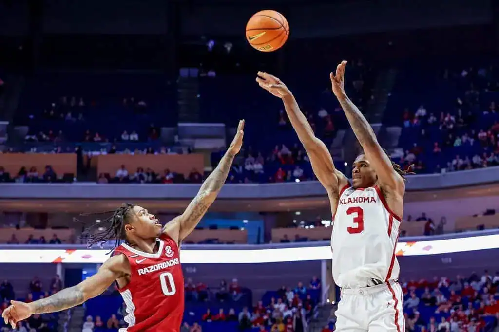 Oklahoma Sooners player taking a jump shot as an Arkansas Razorbacks defender attempts to block it.