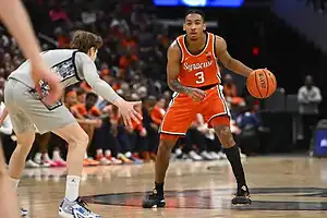 A basketball player in an orange Syracuse uniform dribbles the ball while being closely guarded by an opponent in gray.