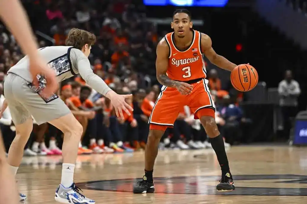 A basketball player in an orange Syracuse uniform dribbles the ball while being closely guarded by an opponent in gray.