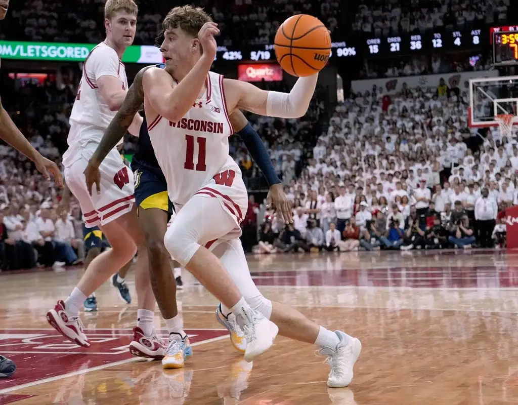 “Wisconsin Badgers #11 driving past a Michigan State Spartans defender in a packed basketball arena.”