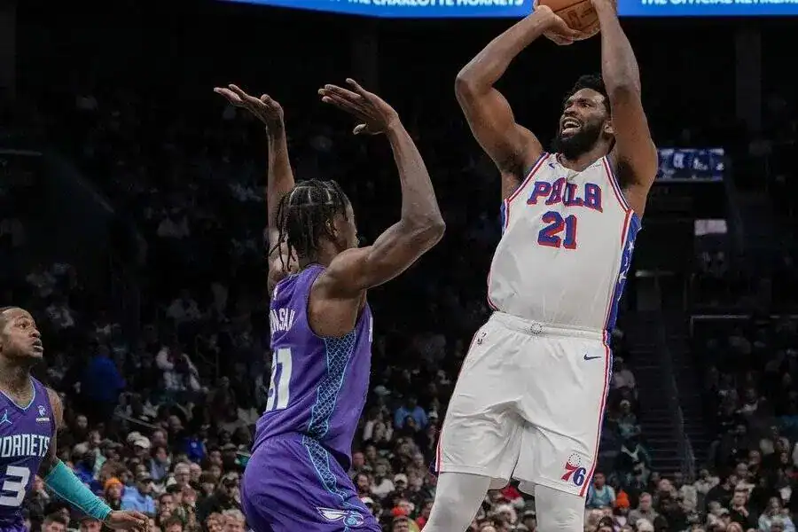 A basketball player in a white Philadelphia 76ers jersey shoots over a defender in a purple Charlotte Hornets uniform.