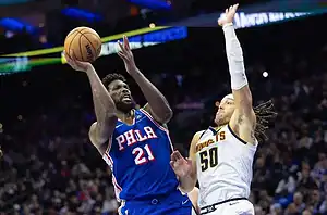 A basketball player in a blue "PHILA" jersey shoots while being defended by an opponent in a white jersey during a game.