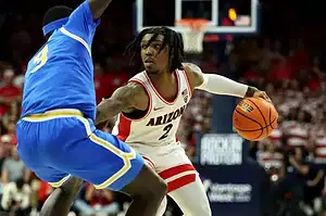 Arizona Wildcats player in a white and red jersey dribbles against a UCLA Bruins defender in a blue uniform.