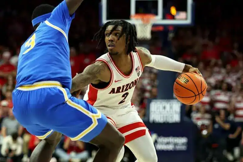 Arizona Wildcats player in a white and red jersey dribbles against a UCLA Bruins defender in a blue uniform.