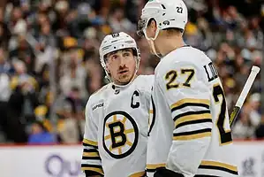 Two Boston Bruins players in white jerseys chat on the ice during a stoppage in play.