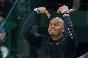 Basketball coach in black raises both arms, signaling while shouting instructions to his team.