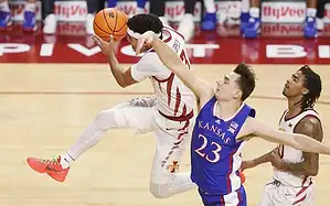 A basketball player leaps in mid-air, preparing to shoot, while another player from the opposing team attempts to block him.