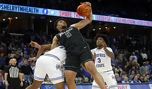 A basketball player in a black jersey leaps to grab a rebound against players in white Memphis State jerseys. The scene is intense and action-packed.