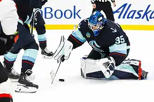 A goalie in a blue hockey uniform crouches on the ice, attempting to catch a puck in a fast-paced game.