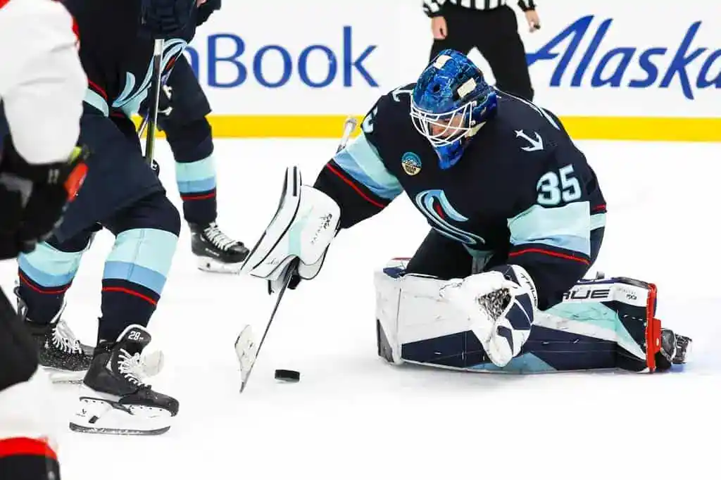 A goalie in a blue hockey uniform crouches on the ice, attempting to catch a puck in a fast-paced game.