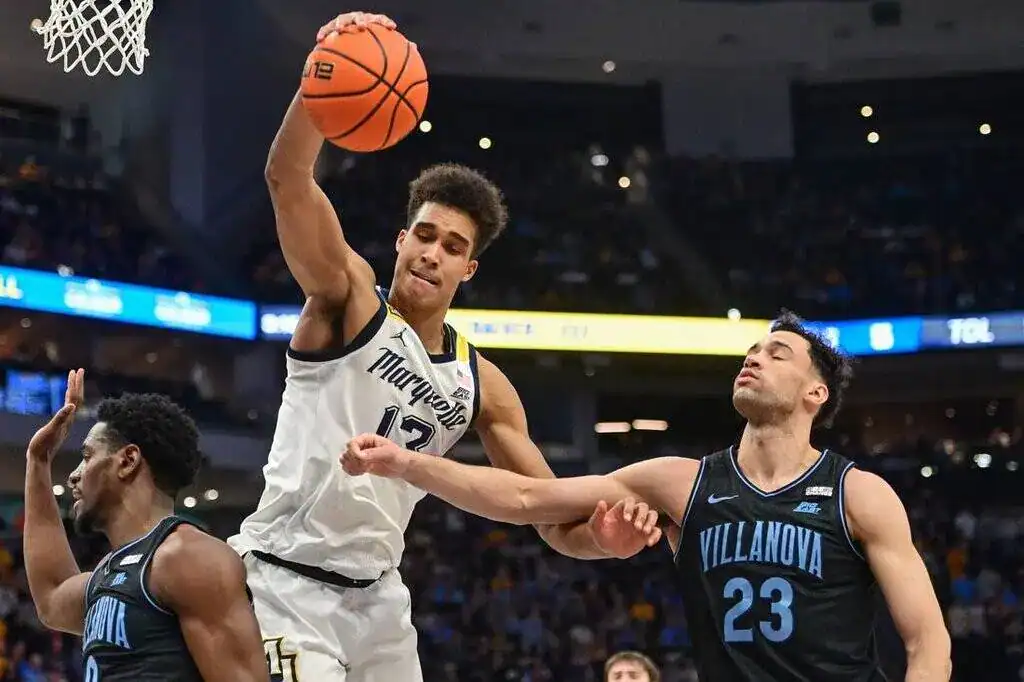 Marquette basketball player goes up for a dunk while a Villanova defender attempts to contest the play during an NCAA game.