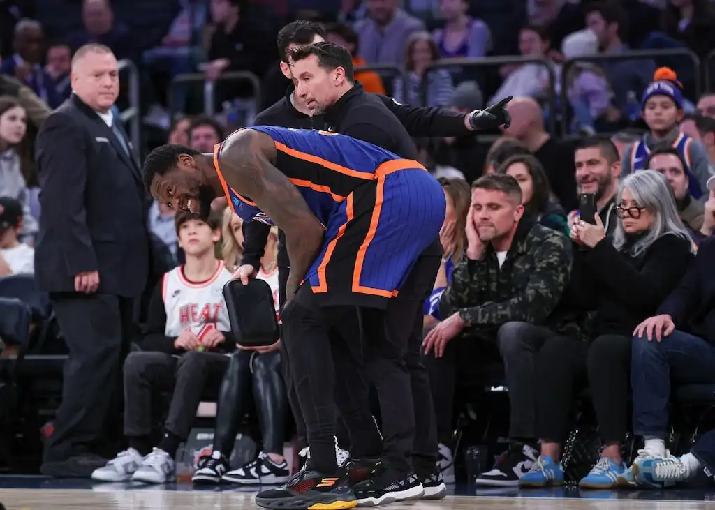 A New York Knicks player grimaces in pain while bending over near the sideline.