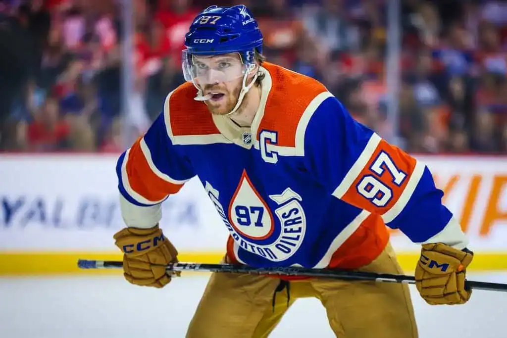 A professional hockey player in an orange and blue Edmonton Oilers uniform, poised on the ice with a determined expression.