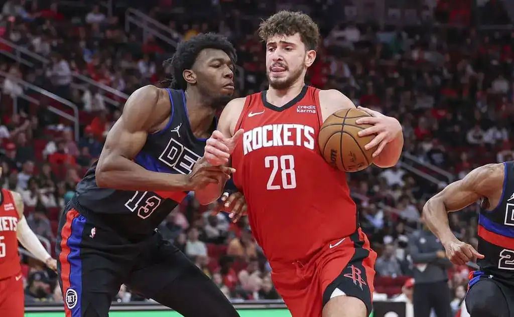 Houston Rockets player handling the ball while being closely defended by a Detroit Pistons player during an NBA game.