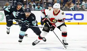 Ottawa Senators forward skates with the puck while pressured by Seattle Kraken defenders during an NHL game.