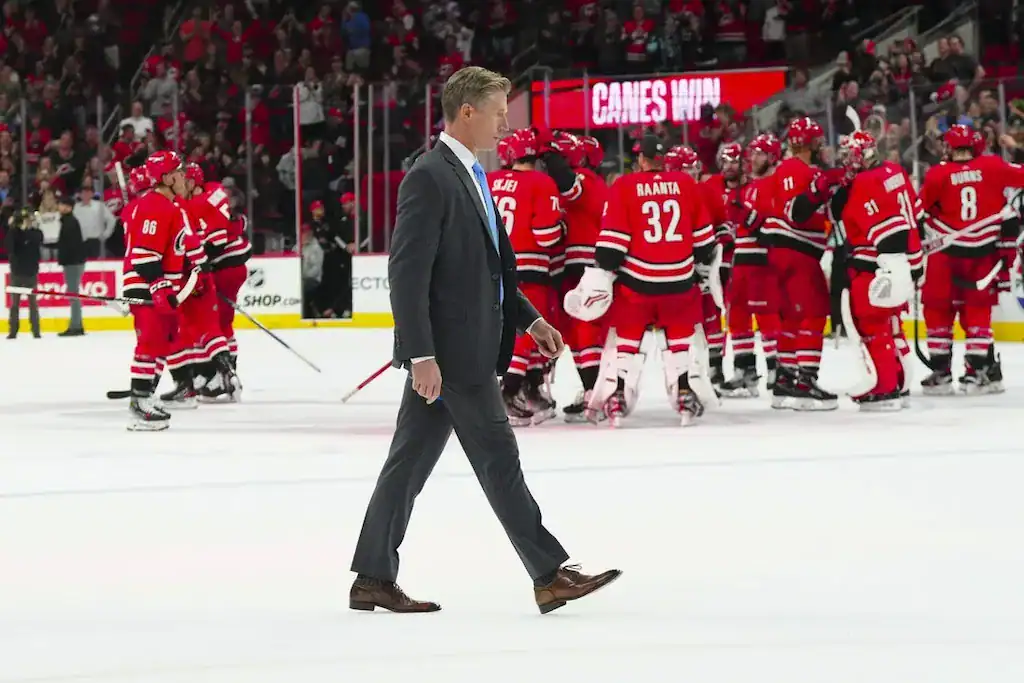 A well-dressed man strides across the ice as Carolina Hurricanes players celebrate a victory in the background, dressed in red jerseys.