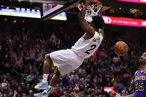 A basketball player hangs from the rim after a dunk, while fans cheer in a packed arena during an intense game.