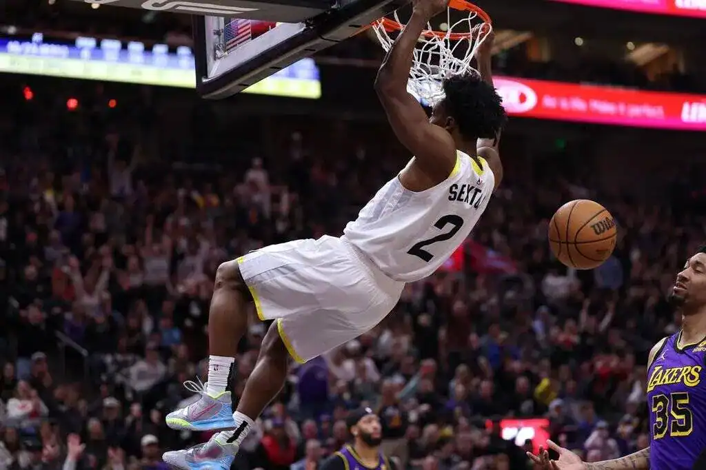 A basketball player hangs from the rim after a dunk, while fans cheer in a packed arena during an intense game.