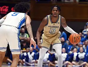 Georgia Tech player in gold uniform dribbles while facing a Duke defender.