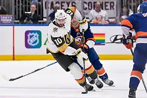 A Vegas Golden Knights player in white skates with the puck while defended by a New York Islanders player.