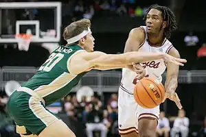 College basketball player in a white uniform passes the ball while being defended by a player in green.