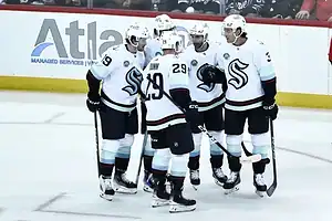 A group of Seattle Kraken players in white jerseys huddle together during an NHL game.