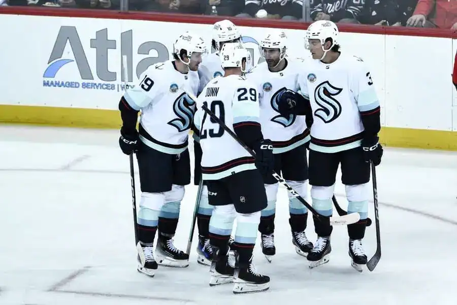 A group of Seattle Kraken players in white jerseys huddle together during an NHL game.