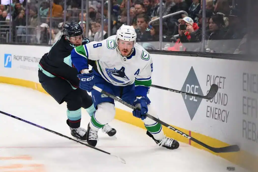 Vancouver Canucks player in a white jersey controls the puck along the boards while being defended.