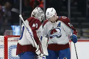 Two Colorado Avalanche hockey players discuss strategy near the goal, with one wearing a goalie mask and the other holding a stick.