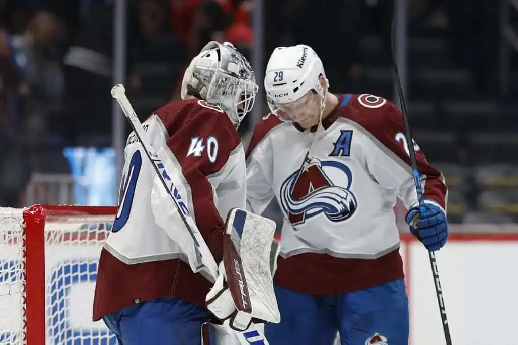 Two Colorado Avalanche hockey players discuss strategy near the goal, with one wearing a goalie mask and the other holding a stick.