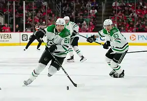 Two Dallas Stars hockey players in white and green uniforms skate on the ice during a game, one handling the puck while the other follows closely behind.