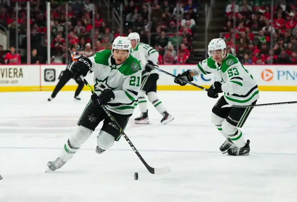 Two Dallas Stars hockey players in white and green uniforms skate on the ice during a game, one handling the puck while the other follows closely behind.
