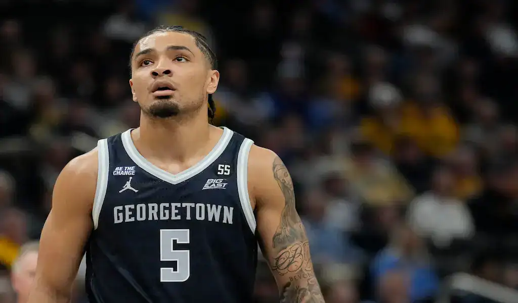 A Georgetown basketball player wearing a navy blue jersey with the number 55, featuring tattoos on his arm, standing on the court with a blurred crowd in the background.