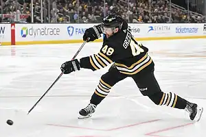 A hockey player in a black and gold uniform takes a powerful slap shot on the ice during a game.