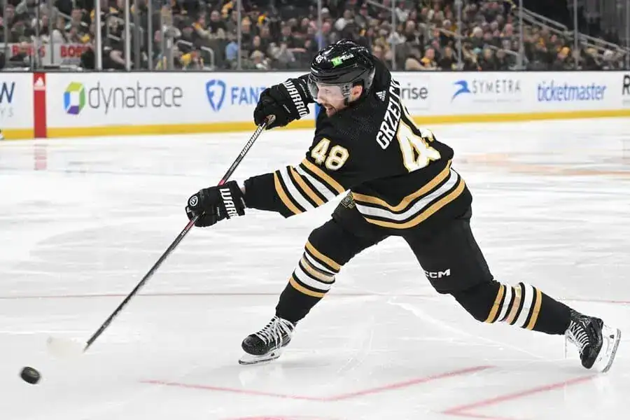 A hockey player in a black and gold uniform takes a powerful slap shot on the ice during a game.