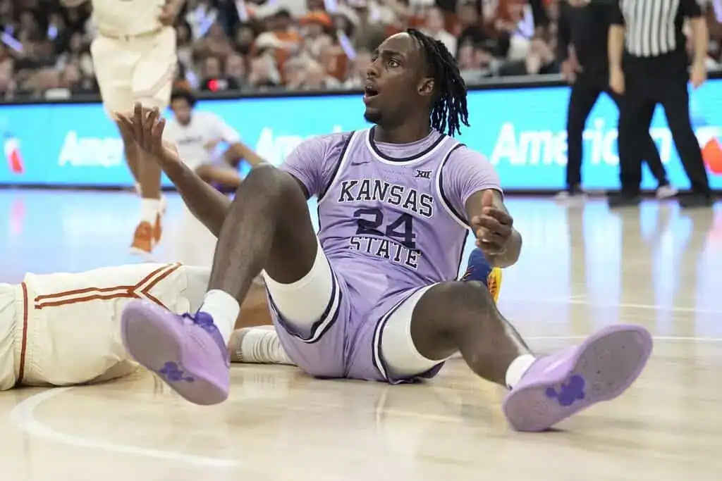 A Kansas State basketball player in a purple jersey sits on the court, showing signs of frustration or defeat after a play.
