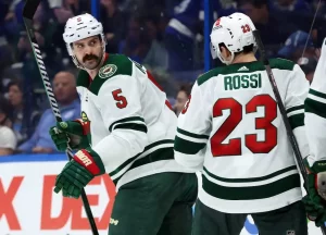 Two Minnesota Wild hockey players in white and green uniforms are seen on the ice during a game.