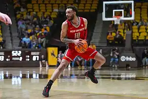 A college basketball player in a red New Mexico jersey dribbles and prepares to drive toward the basket.