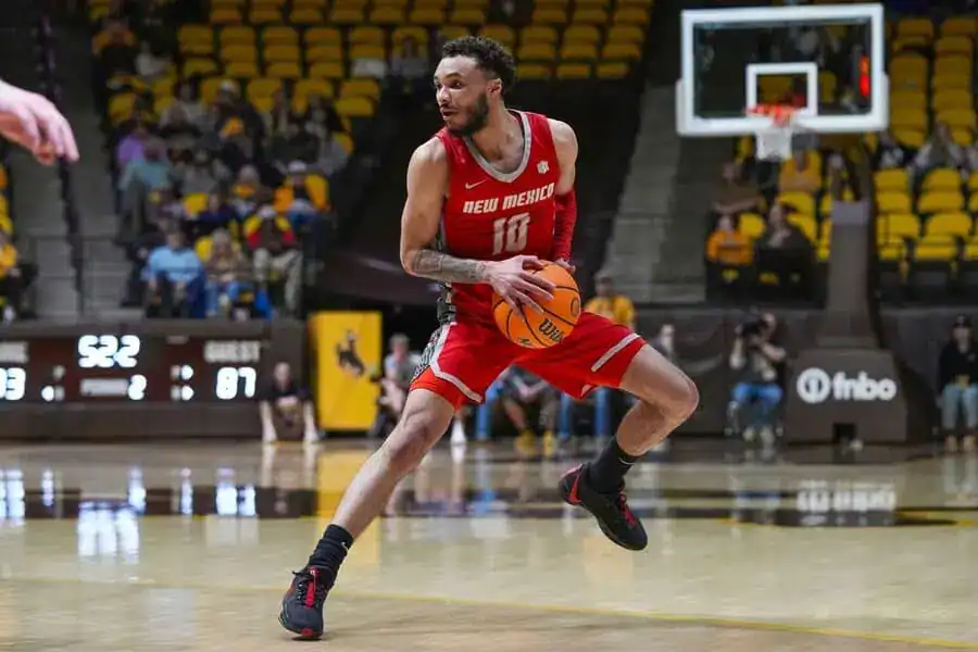 A college basketball player in a red New Mexico jersey dribbles and prepares to drive toward the basket.