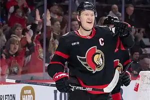 A hockey player in a black jersey with a red logo celebrates a goal, surrounded by cheering fans in a lively arena.